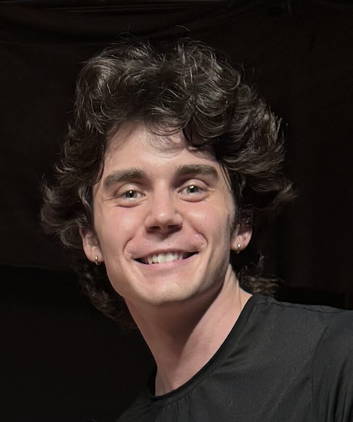A dark haired Caucasian man with normal eyebrows, smiling in front of a black background